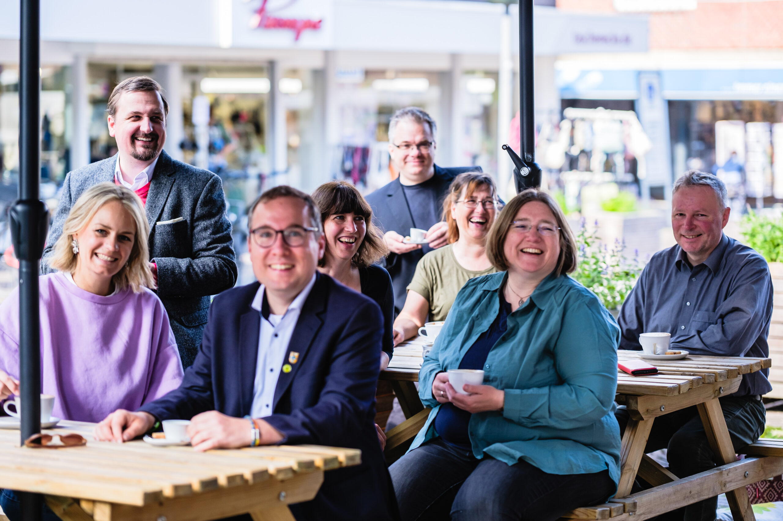 Acht Personen sitzen oder stehen lachend an zwei hölzernen Picknicktischen im Außenbereich eines Cafés in der Dülmener Innenstadt. Sie halten weiße Kaffeetassen in den Händen. Im Hintergrund sind Ladenfronten und ein Sonnenschirm zu sehen. Die Gruppe trägt Alltagskleidung wie Sakkos, Blusen & Pullover.