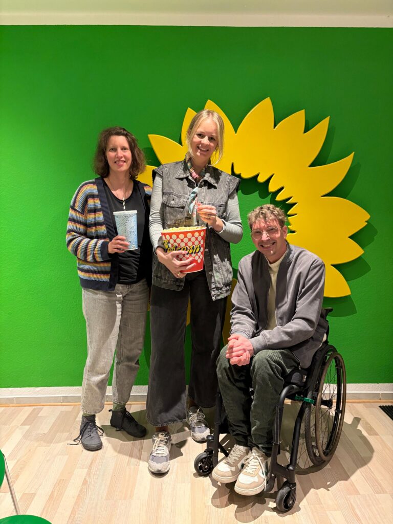 Drei lächelnde Personen posieren vor einer grünen Wand mit dem gelben Sonnenblumen-Logo der Grünen. Eine Frau links trägt eine bunt gestreifte Strickjacke, die Frau in der Mitte hält einen rot-weiß gepunkteten Popcorn-Eimer und Dennis Sonne sitzt rechts im Rollstuhl. Alle drei blicken direkt in die Kamera; im Vordergrund ist ein heller Holzboden sichtbar.