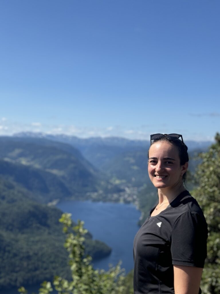 Eine weiblich gelesene Frau schaut in die Kamera und im Hintergrund ein Tal mit See