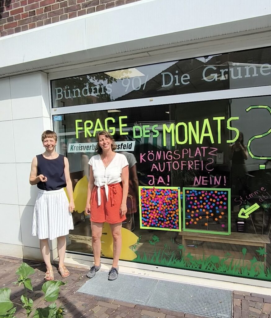Zwei lächelnde Frauen stehen vor dem Schaufenster des Grünen-Büros in Dülmen. Auf der Scheibe ist die Frage „Königsplatz autofrei?“ mit zwei Abstimmungsfeldern für „Ja!“ und „Nein!“ zu sehen, beide gefüllt mit bunten Punkten.